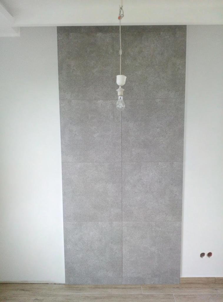 Pionowy panel z szarych płytek imitujących beton, zamontowany na ścianie pomiędzy dwiema białymi ścianami, z wiszącą żarówką na kablu i widocznymi otworami na gniazdka elektryczne.