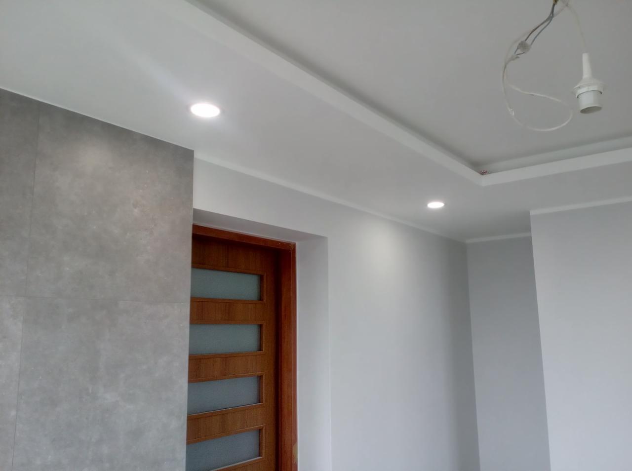 Wykończone wnętrze: szara ściana z płytkami imitującymi beton, drewniane drzwi z przeszkleniami, biały sufit z oświetleniem punktowym i listwami dekoracyjnymi.