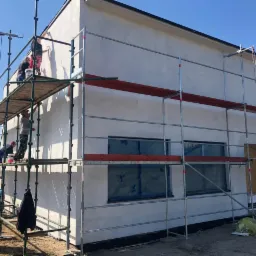 Ekipa budowlana na rusztowaniu podczas nakładania jasnej elewacji na dom jednorodzinny, widoczne okna zabezpieczone folią i narzędzia pracy.