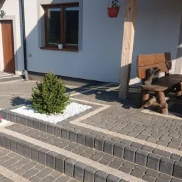 Ułożona kostka brukowa w odcieniach szarości z dekoracyjnymi elementami z białego kamienia i roślinności przed domem z drewnianymi oknami i meblami ogrodowymi.