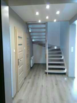 Jasne, drewniane schody z podświetleniem LED prowadzące na górę, widoczne z dołu klatki schodowej. Schody proste i zabiegowe. Na ścianie widoczne drzwi i grzejnik.