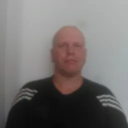 Portret mężczyzny w czarnej koszulce z trzema białymi paskami na tle jasnej ściany, wykonany w Gromadce. Ujęcie od przodu, skupione na twarzy.