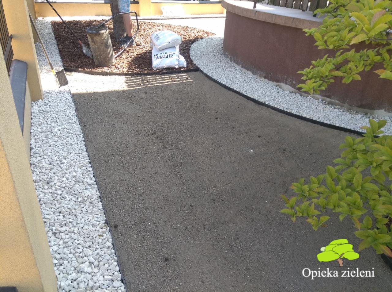 Przygotowanie podłoża pod trawnik z rolki: wyrównana ziemia, obrzeża z białych kamyków, kora wokół drzewa, worki z ziemią i narzędzia w tle.