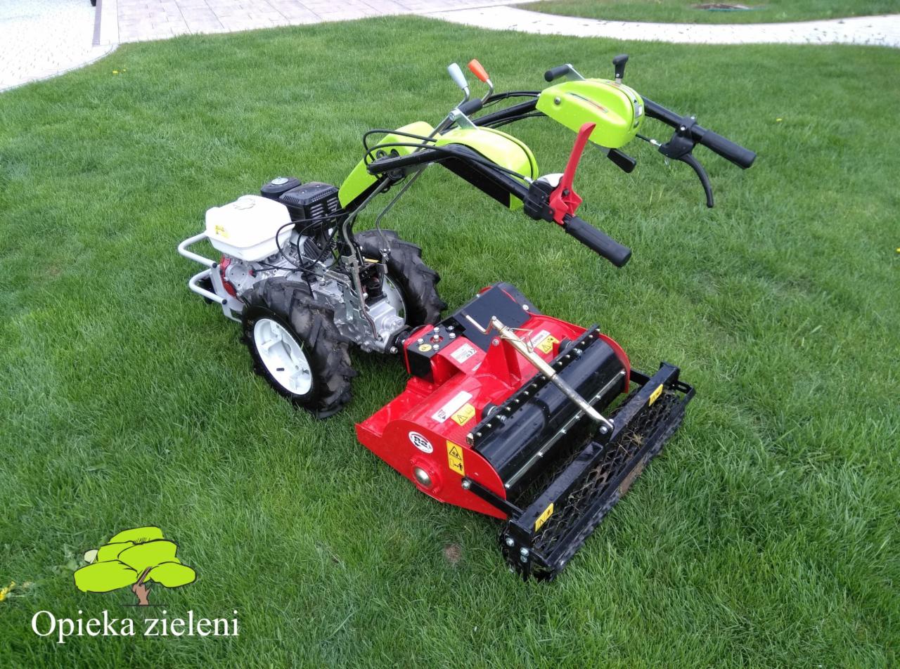 Profesjonalny aerator spalinowy z zielonymi elementami na zadbanym trawniku, widoczne ostrzeżenia bezpieczeństwa na urządzeniu.