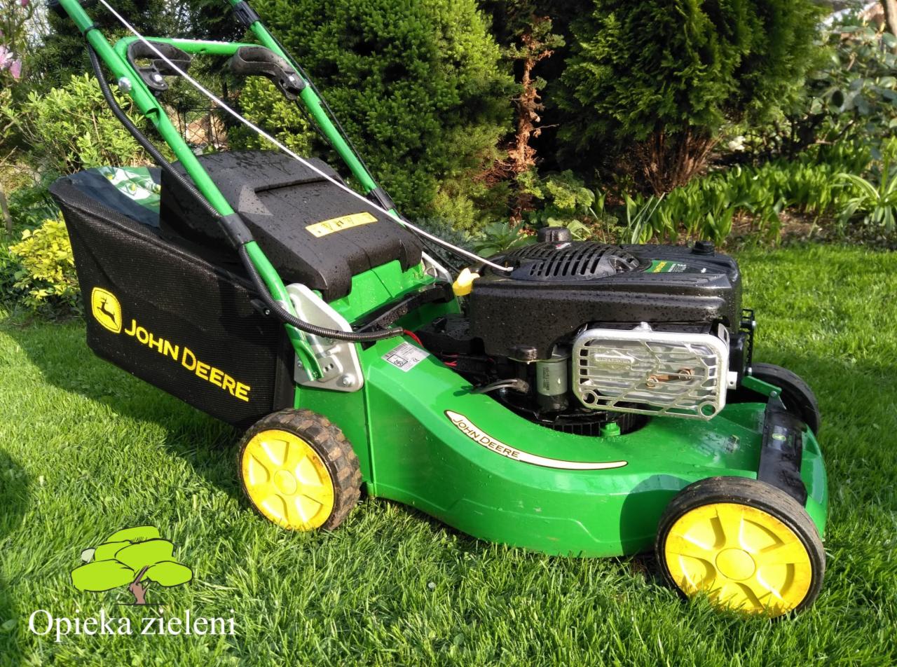 Zielona kosiarka spalinowa John Deere na trawniku, z widocznym koszem na trawę i elementami silnika, w tle krzewy i drzewa.