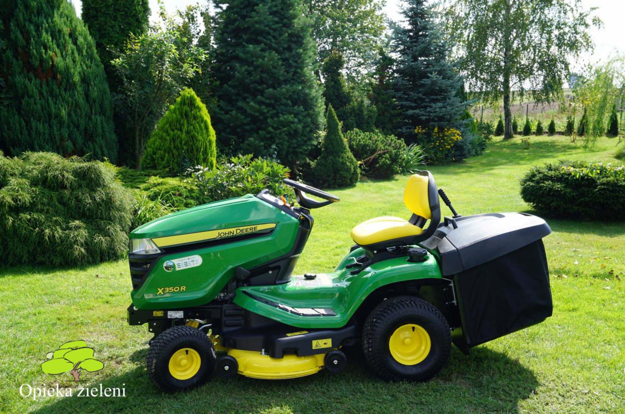 Zielony traktorek kosiarka John Deere X350R z żółtym siedzeniem na trawniku, w tle różnorodne drzewa i krzewy ozdobne, widoczny kosz na trawę.