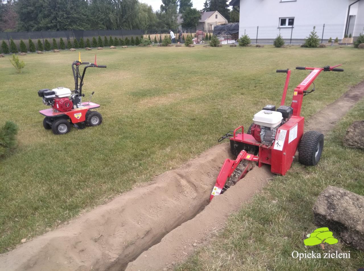 Czerwona glebogryzarka spalinowa w trakcie wykopywania wąskiego rowu w trawniku, obok druga mniejsza glebogryzarka, w tle budynek mieszkalny i rząd tuj.
