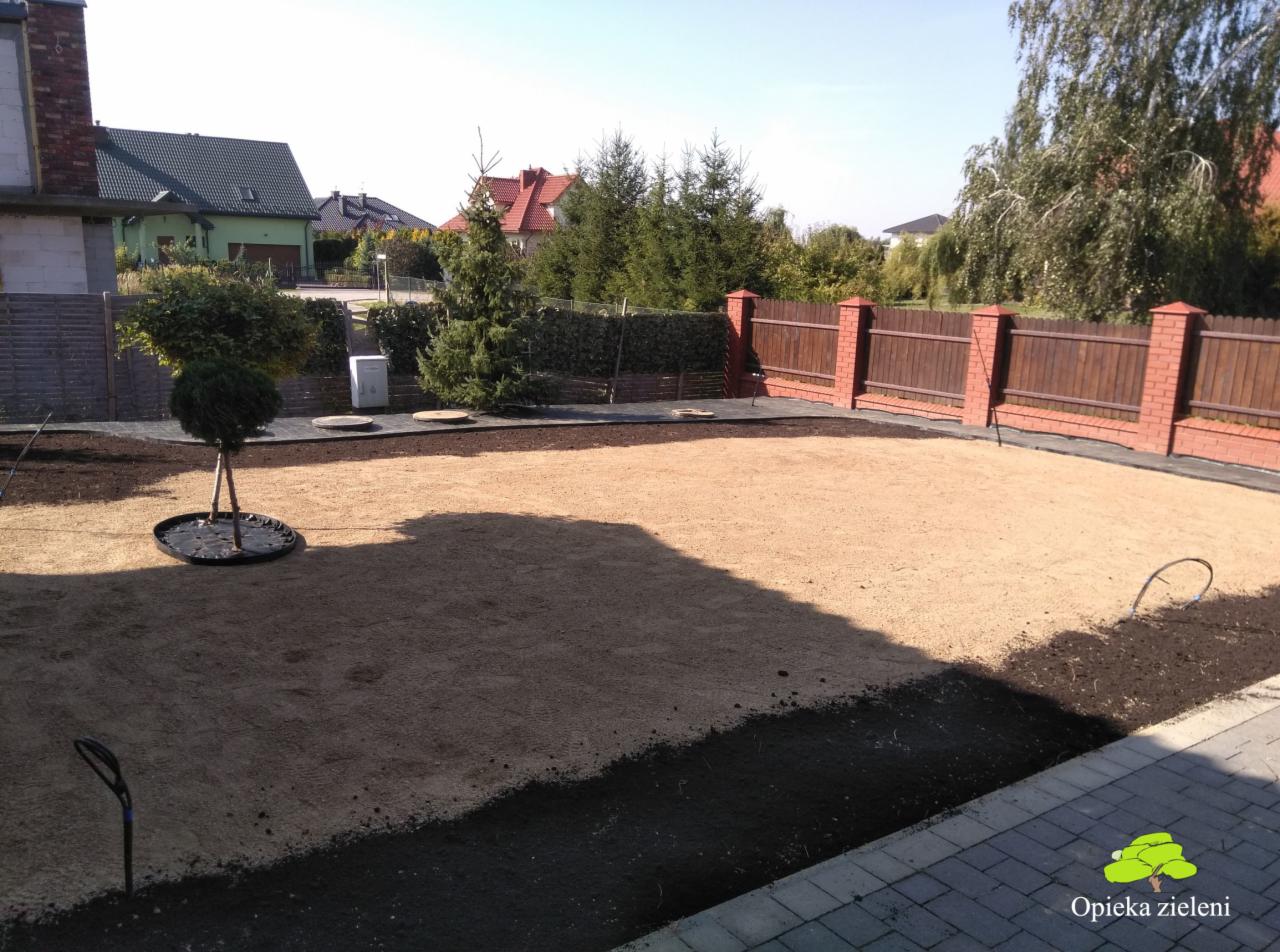 Przygotowany teren pod trawnik, piaskowanie, widoczny system nawadniania, obrzeża z ciemnej ziemi, w tle ogrodzenie z ceglanymi słupkami i drewnianymi panelami, na pierwszym planie kostka brukowa.