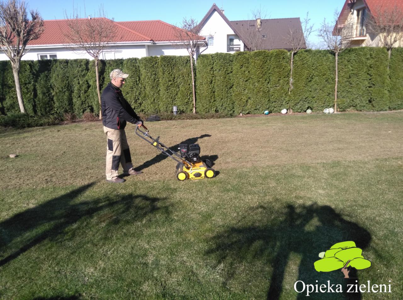 Mężczyzna w Lublinie używa żółtej wertykulatora na trawniku przed żywopłotem; w tle domy.