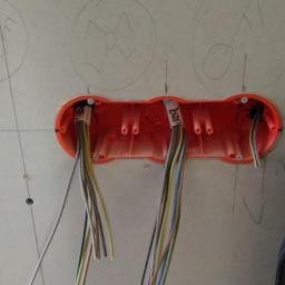 Rsinformatyka - Pomarańczowa puszka elektryczna z przewodami wystającymi ze ściany gipsowo-kartonowej z naniesionymi odręcznymi oznaczeniami, widoczny fragment odzieży roboczej.