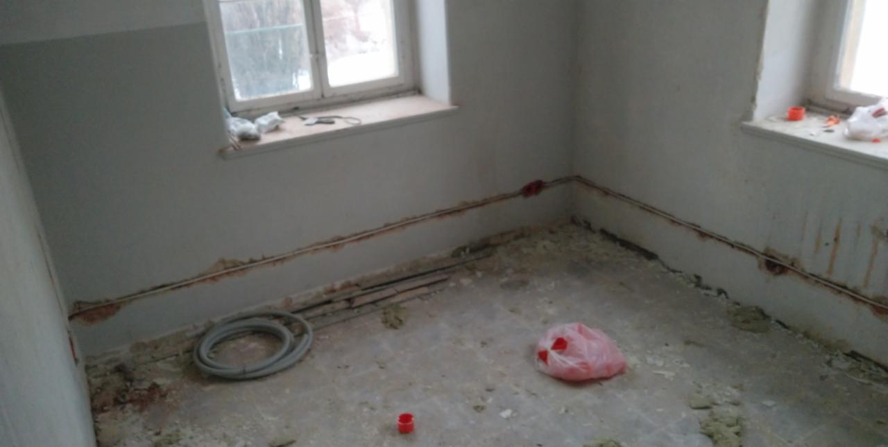 Pokój w trakcie remontu, z widocznymi przewodami elektrycznymi w bruzdach na ścianach, zwiniętym kablem na podłodze oraz resztkami gruzu i plastikową torbą.