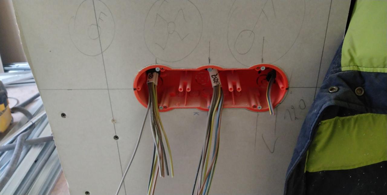 Pomarańczowa puszka elektryczna z przewodami wystającymi ze ściany gipsowo-kartonowej z naniesionymi odręcznymi oznaczeniami, widoczny fragment odzieży roboczej.