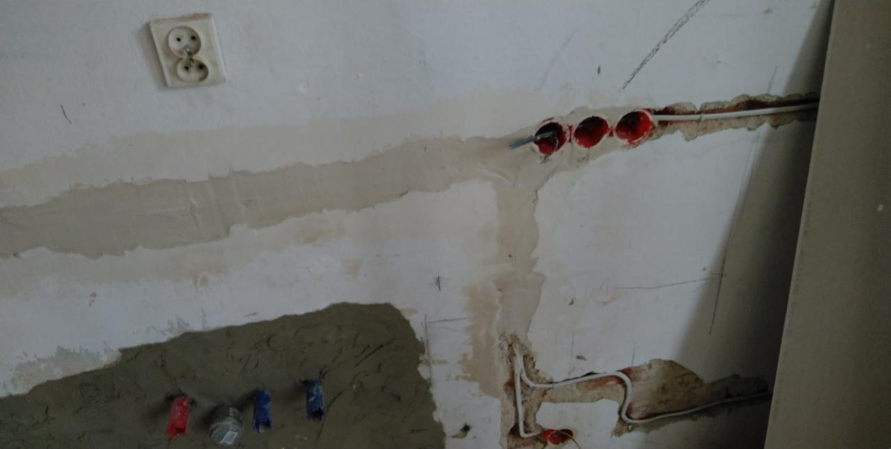 Ściana w trakcie remontu z widocznymi przewodami elektrycznymi w czerwonych puszkach, gniazdko elektryczne, oraz fragmenty ściany pokryte szpachlą i świeżym tynkiem.