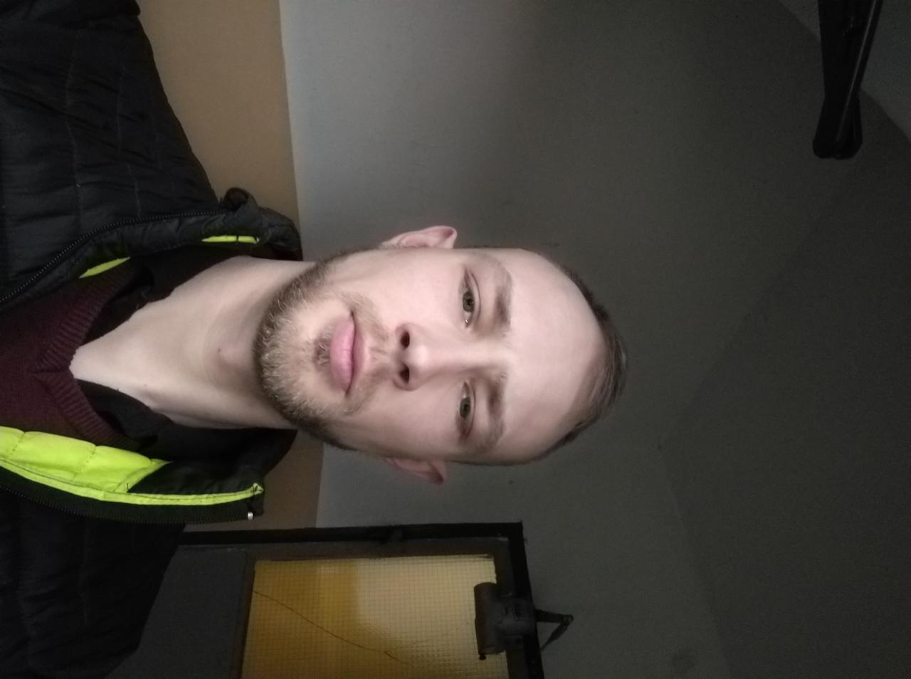 Portret mężczyzny z brodą, ubranego w kurtkę z neonowymi elementami, w pomieszczeniu z oknem. Nietypowy kadr z dołu.