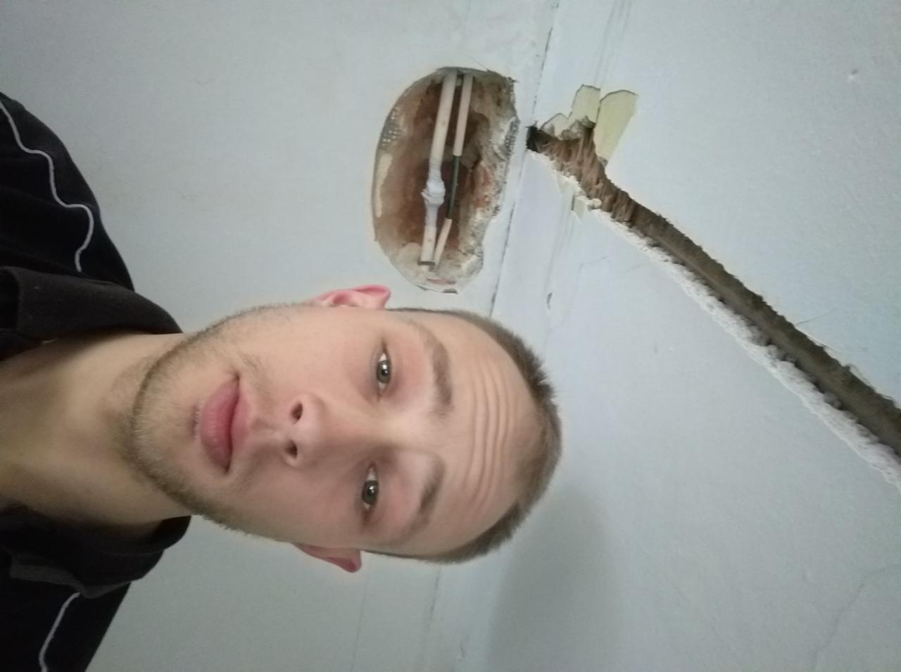 Autoportret elektryka na tle wyżłobienia w ścianie z wystającymi kablami i puszką elektryczną w trakcie prac instalacyjnych.