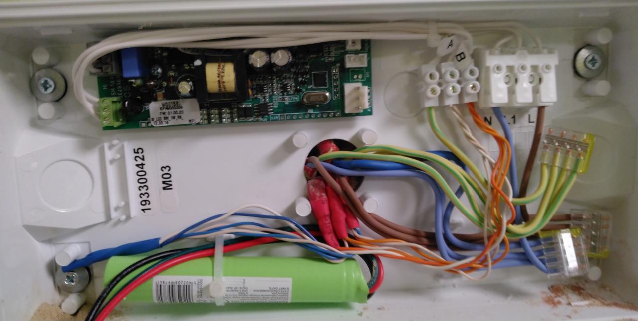 Wnętrze białej puszki elektrycznej z podłączonymi przewodami o różnych kolorach, widoczna płytka drukowana, złącza i akumulator. Przewody połączone złączkami Wago.