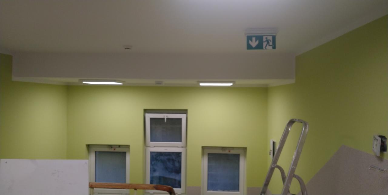 Wnętrze pomalowane na jasnozielony kolor z trzema oknami, aluminiową drabiną i oświetleniem sufitowym, w tym znak ewakuacyjny.