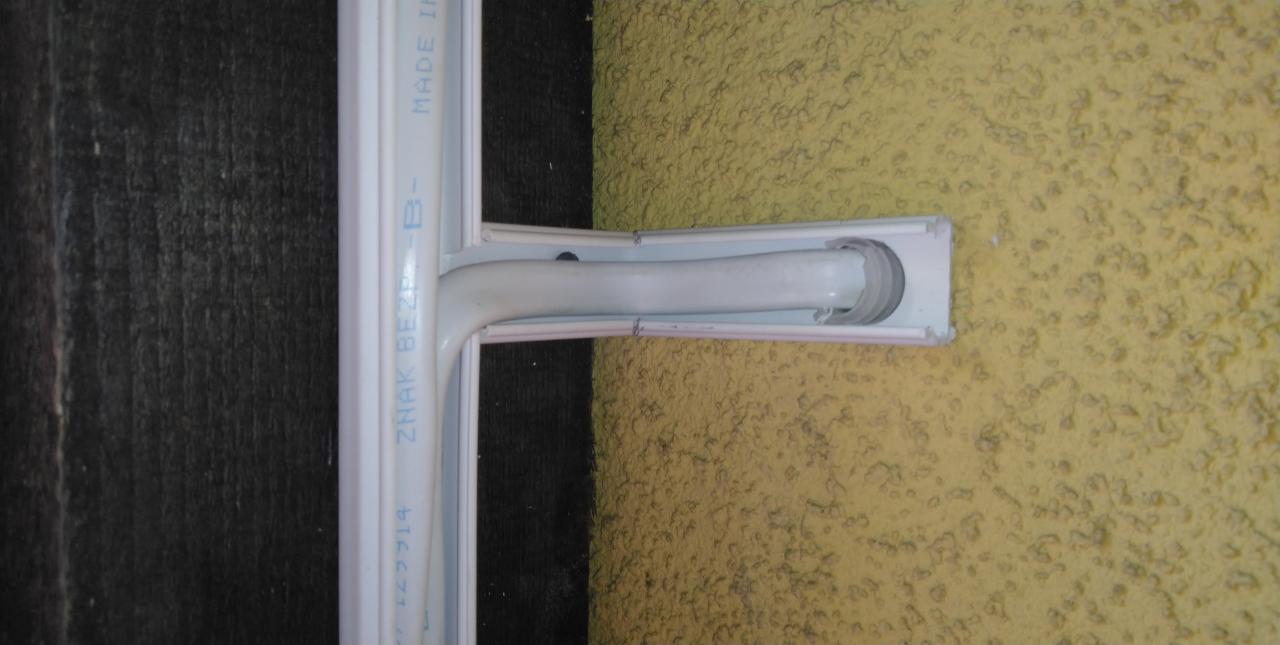 Fragment okna PCV z widocznym napisem 'Znak Bezpieczeństwa', zamontowanego na żółtej, chropowatej ścianie.