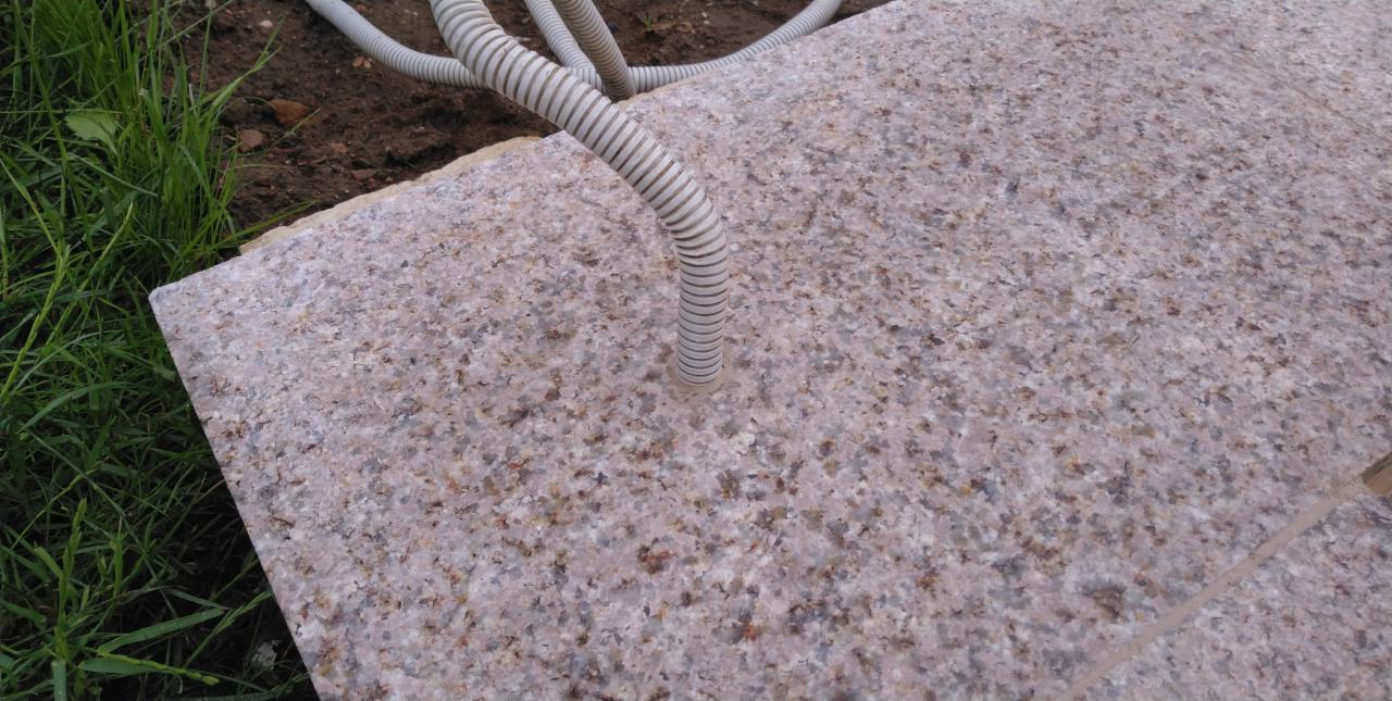 Szara, karbowana rura wychodząca z granitowej płyty w Boguszowie-Gorcach, otoczona zieloną trawą.