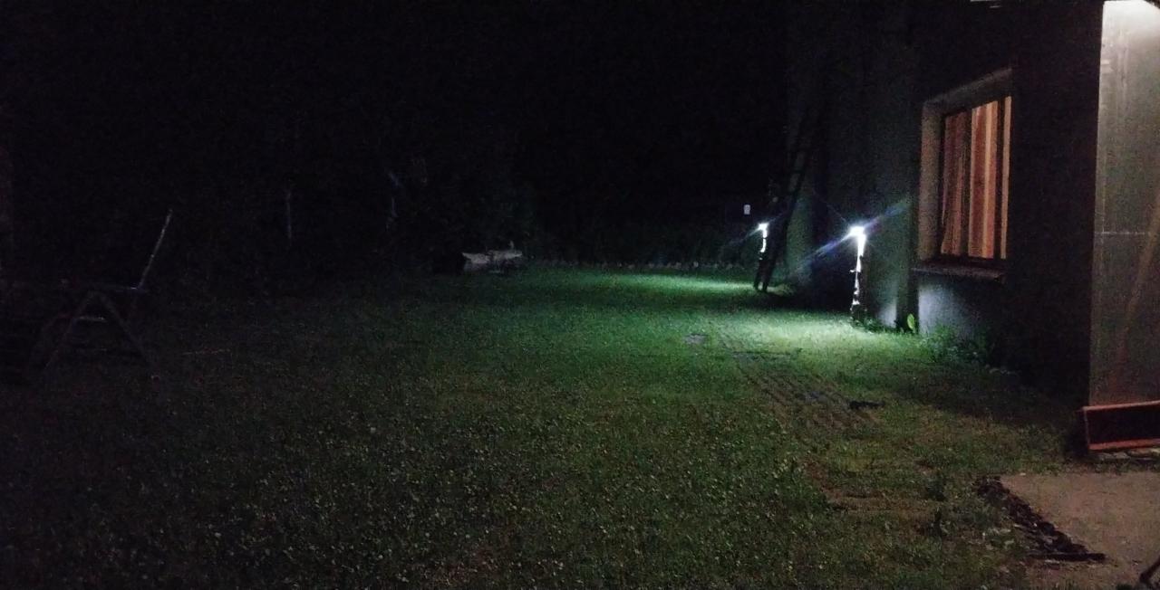 Oświetlony trawnik przed domem nocą, widoczne lampy ogrodowe i okno z zasłoniętymi zasłonami.