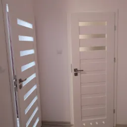 Dwa białe, nowoczesne drzwi wewnętrzne z przeszkleniami i poziomymi panelami, jeden otwarty, drugi zamknięty, zamontowane w jasnym pomieszczeniu na podłodze z paneli.