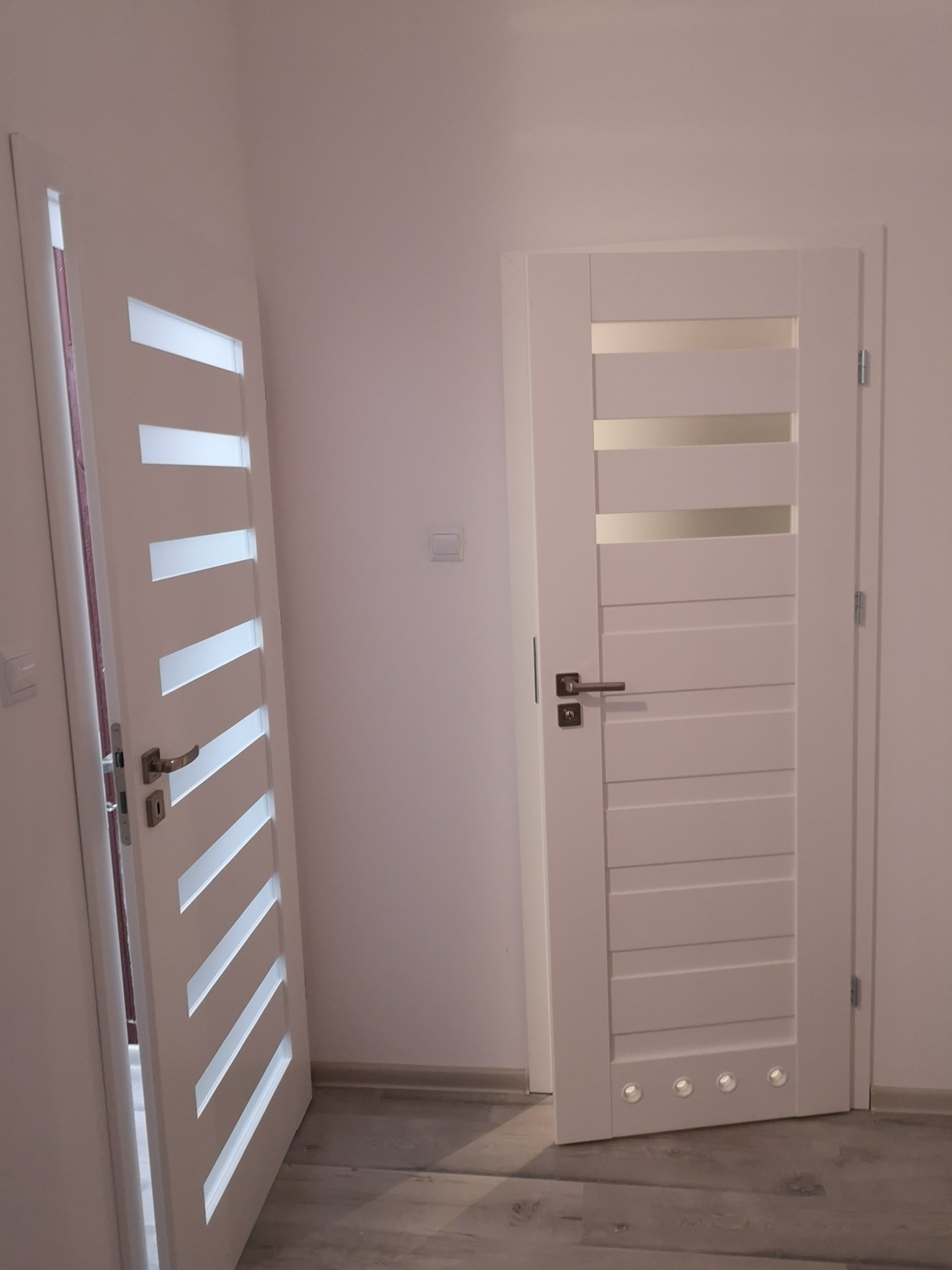 Dwa białe, nowoczesne drzwi wewnętrzne z przeszkleniami i poziomymi panelami, jeden otwarty, drugi zamknięty, zamontowane w jasnym pomieszczeniu na podłodze z paneli.