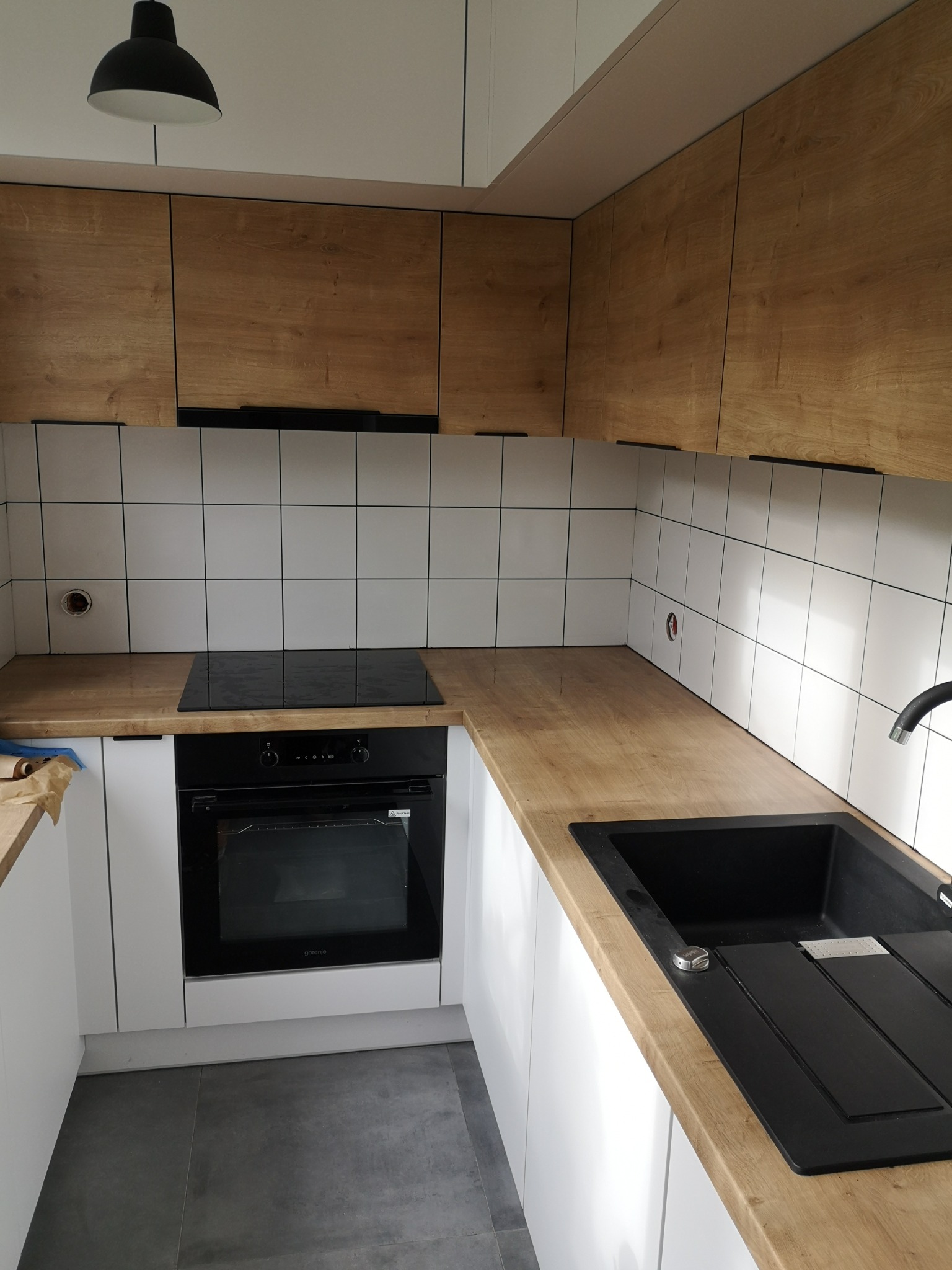 Jasna kuchnia z drewnianymi szafkami górnymi, białymi szafkami dolnymi, czarnym zlewem i płytą indukcyjną, białe kwadratowe płytki na ścianie, szare płytki podłogowe, czarna lampa sufitowa.