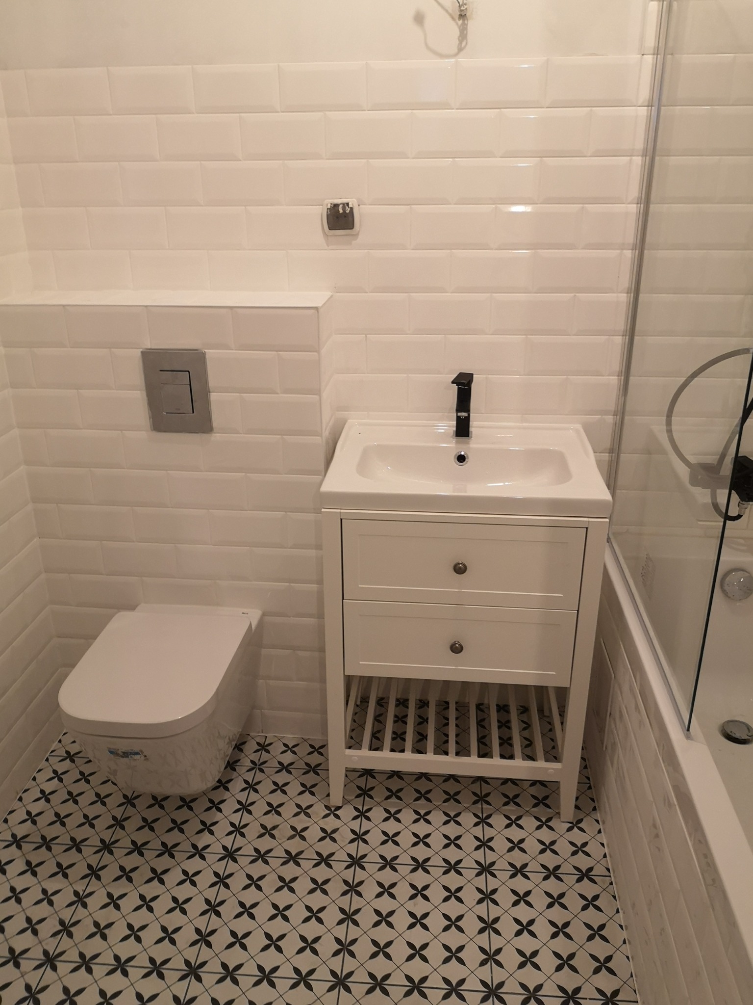 Nowoczesna łazienka z białymi kafelkami typu metro, umywalką na szafce z szufladami, toaletą podwieszaną i wzorzystą podłogą w odcieniach bieli i czerni.