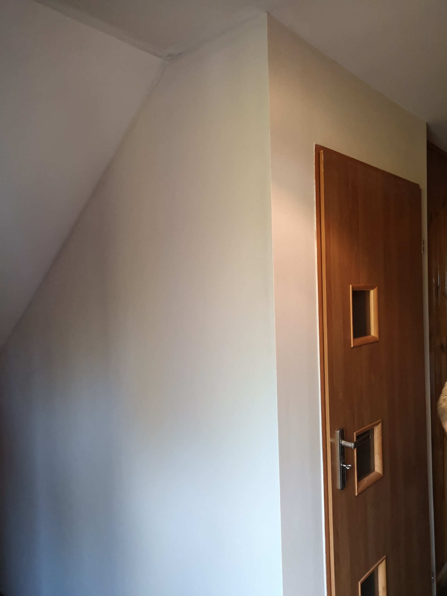 Ściana po malowaniu w odcieniach bieli, widoczna drewniana ościeżnica drzwi z małymi okienkami i klamką.