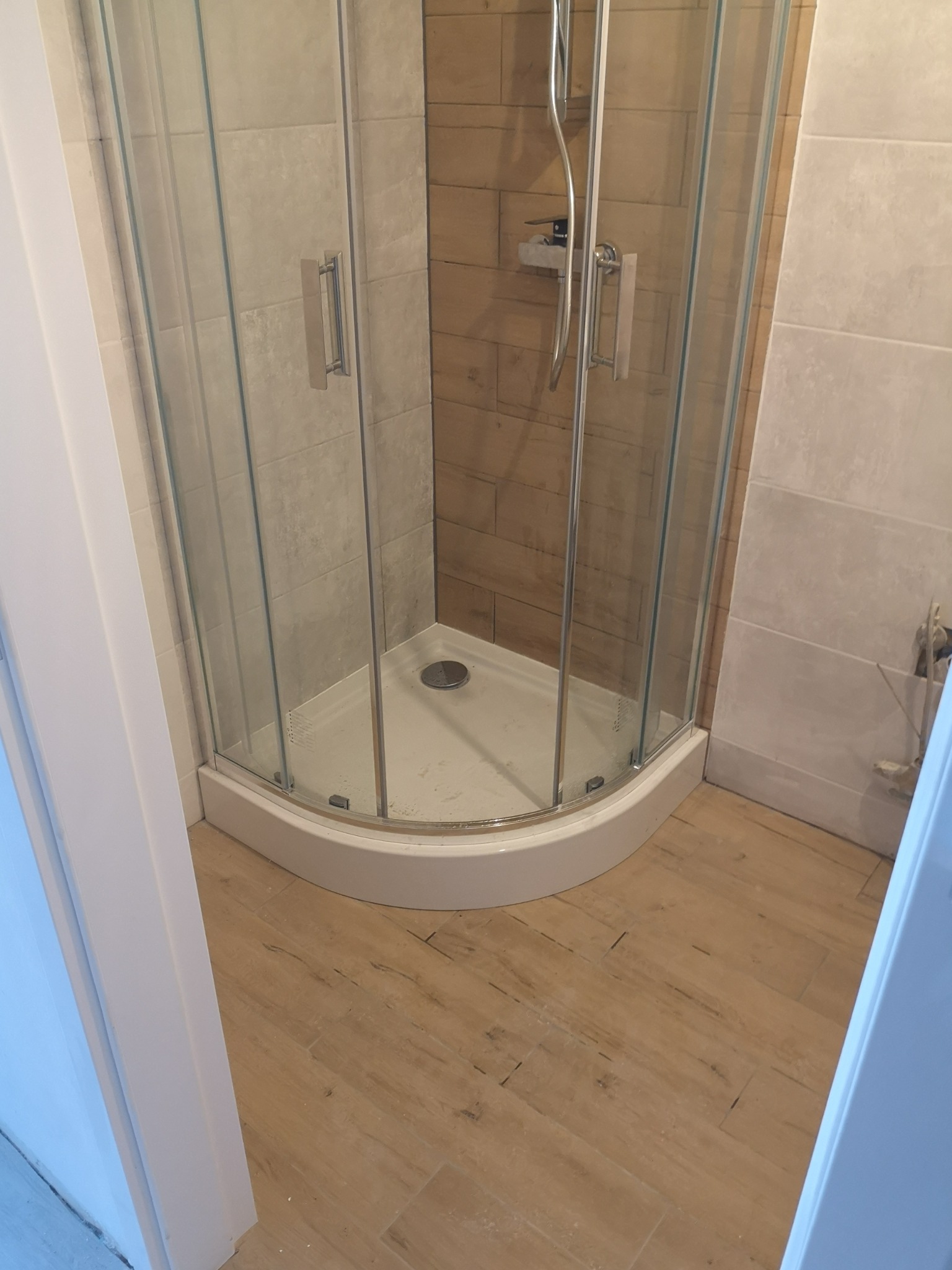 Narożna kabina prysznicowa z chromowanymi uchwytami i armaturą, widoczna na tle płytek imitujących drewno i jasnych płytek ściennych