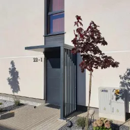 Nowoczesne zadaszenie nad wejściem do domu z metalową konstrukcją, numerem 22-1 i młodym drzewkiem o bordowych liściach.