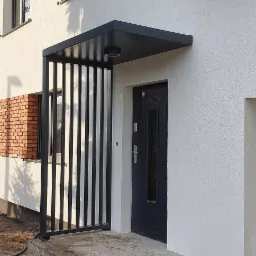 Nowoczesne zadaszenie nad wejściem do domu z pionowymi, czarnymi elementami, oświetleniem punktowym i ciemnymi drzwiami wejściowymi, przylegające do ściany z ceglanym fragmentem.
