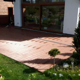 Nowoczesny taras kompozytowy w kolorze ciepłego brązu, przylegający do domu z dużym oknem przesuwnym, otoczony zielonym trawnikiem z ozdobnymi krzewami i drzewkami.