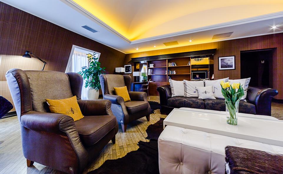 Salon wypoczynkowy z brązowymi fotelami uszakami, sofą z poduszkami z motywem nut, białym pufem i żółtymi tulipanami w wazonie na niskim stoliku, oświetlony ciepłym światłem.