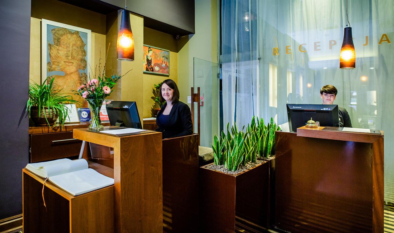 Recepcja hotelowa z dwoma pracownikami za drewnianymi ladami, widoczne rośliny doniczkowe, wiszące lampy i elementy dekoracyjne, napis 'Recepcja' na szklanej ścianie w tle.