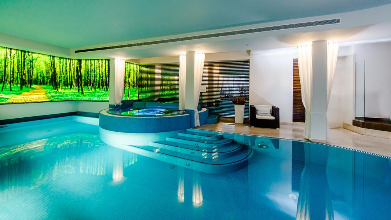 Luksusowy kryty basen z jacuzzi, podświetlony obraz lasu na ścianie, designerskie zasłony i eleganckie meble.