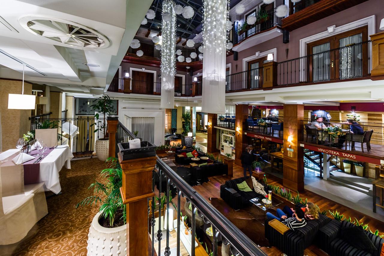 Wielopoziomowe wnętrze hotelu z restauracją Vinoteka Tapas, widok z góry na stoliki, sofy, dekoracje świetlne i balkony z kwiatami.