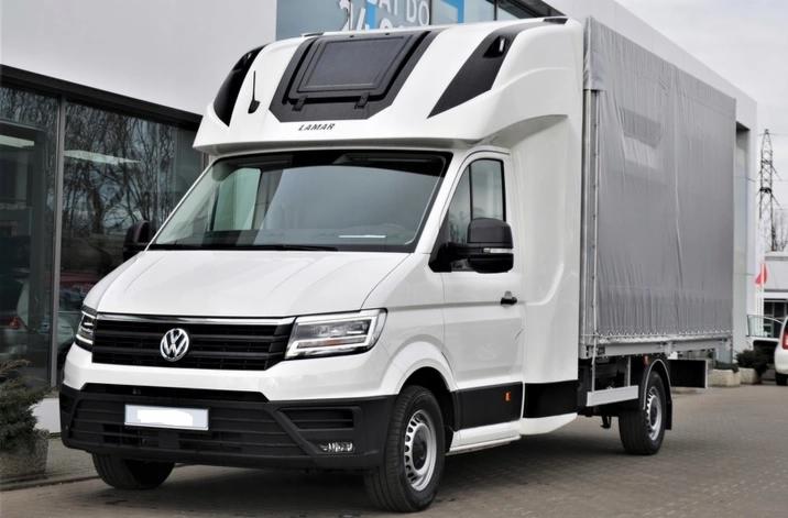 Biały samochód dostawczy marki Volkswagen z zabudową plandekową i napisem LAMAR na kabinie, zaparkowany przed budynkiem z przeszkloną fasadą.