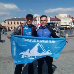 Dwóch mężczyzn trzyma niebieski baner z napisem TOURSport RUNNING TEAM i logiem górskim na tle miejskiego placu z budynkami w tle.
