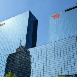 Dwa nowoczesne szklane wieżowce, na jednym widoczne logo firmy ubezpieczeniowej, na drugim odbicie innego budynku, błękitne niebo w tle.