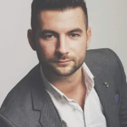 Portret mężczyzny w szarej marynarce i białej koszuli, z delikatnym zarostem i ciemnymi włosami zaczesanymi do tyłu, na jasnym tle.