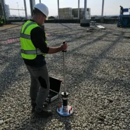 Pracownik w kasku i kamizelce odblaskowej używa dynamicznego obciążenia płytowego do badania zagęszczenia gruntu na placu budowy z widoczną konstrukcją stalową w tle.