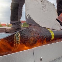 Kocie Ruchy Oskar Klimaszewski - Dekarz topi papę na dachu za pomocą palnika. Widoczne płomienie, stopiona papa i fragment narzędzi. W tle zabudowa jednorodzinna.