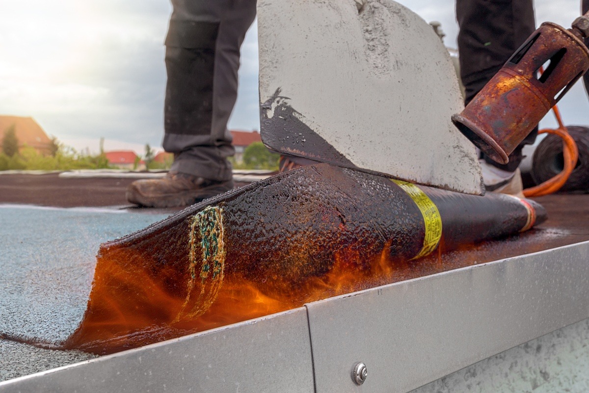 Dekarz topi papę na dachu za pomocą palnika. Widoczne płomienie, stopiona papa i fragment narzędzi. W tle zabudowa jednorodzinna.