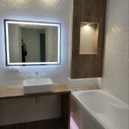 Nowoczesna łazienka z podświetlanym lustrem, umywalką nablatową na drewnianym blacie, wanną z podświetleniem LED i dekoracyjnymi białymi płytkami 3D oraz wnęką z drewnopodobnymi płytkami.