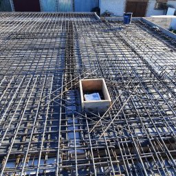 Paśnik - Zbrojenie pod wylewkę na przygotowanym szalunku, widoczna siatka z prętów stalowych i drewniana skrzynka z dokumentami w rogu. W tle budynek gospodarczy i pole.