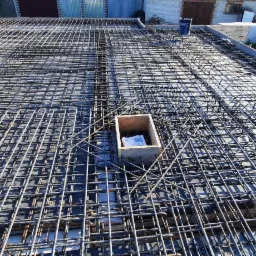 Zbrojenie pod wylewkę na przygotowanym szalunku, widoczna siatka z prętów stalowych i drewniana skrzynka z dokumentami w rogu. W tle budynek gospodarczy i pole.