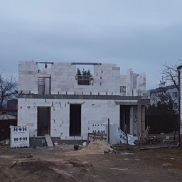 Paśnik - Surowy stan domu w trakcie budowy, ściany z białych bloczków, widoczne otwory okienne i drzwiowe, materiały budowlane H+H na placu budowy, pochmurne niebo w tle.