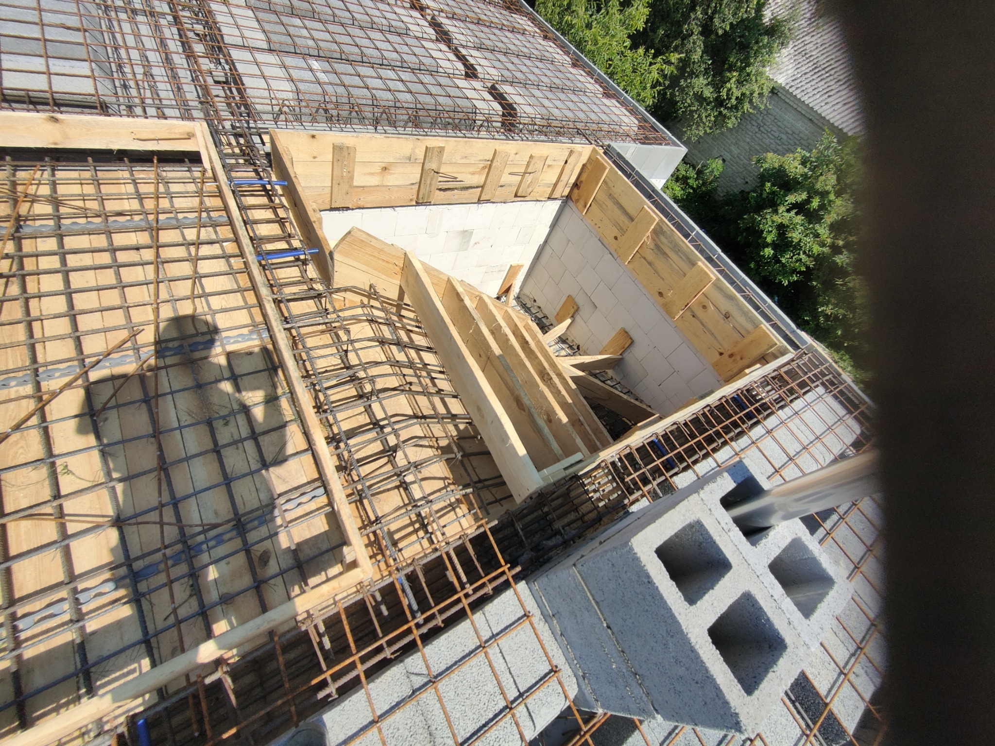 Wiązanie zbrojenia stropu na drewnianym szalunku, widoczny cień osoby robiącej zdjęcie, bloczki betonowe i elementy murowane.