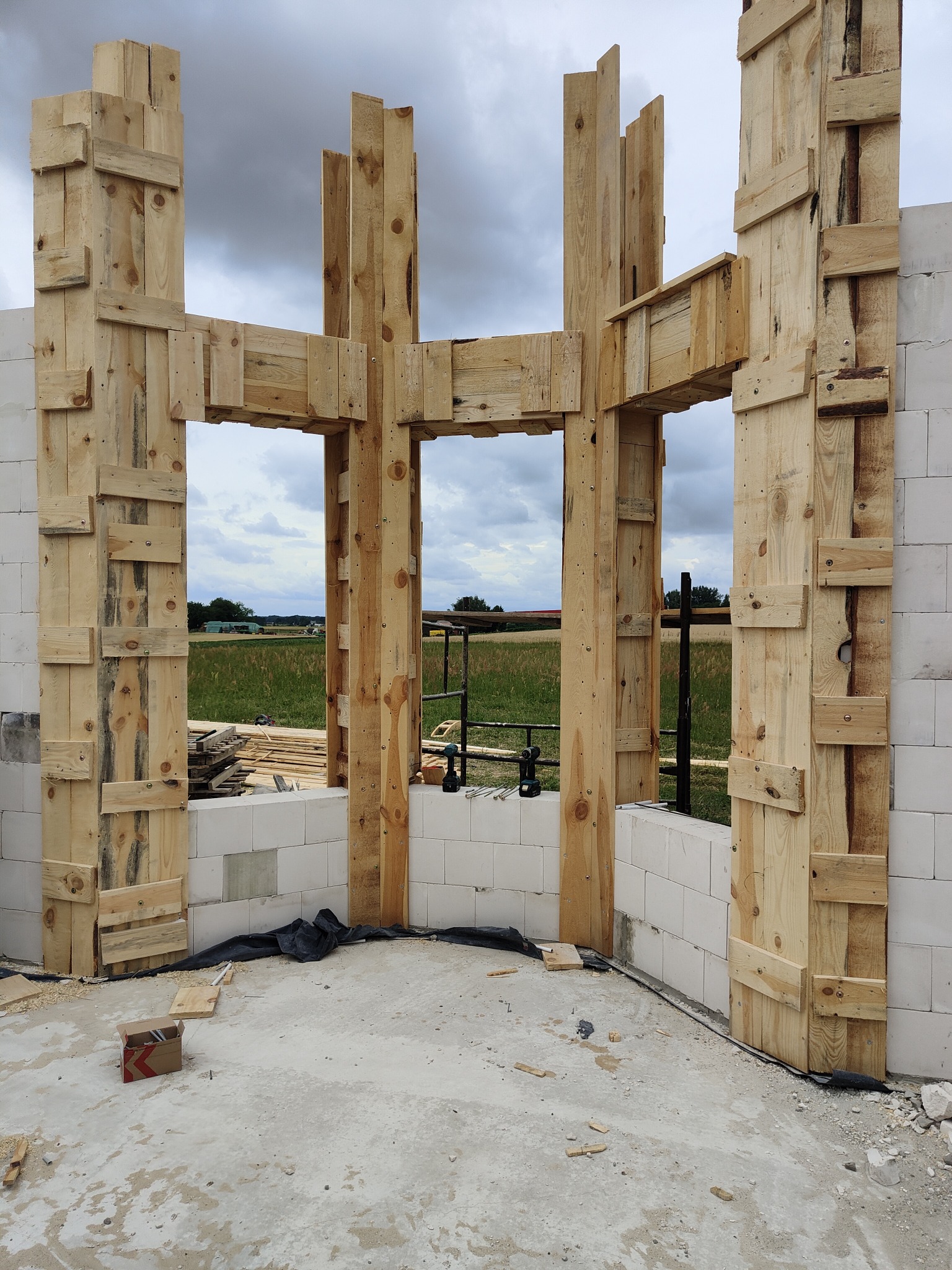 Wnętrze w trakcie budowy, widoczne ściany z białych bloczków i drewniana konstrukcja szalunkowa okien, z widokiem na pola i pochmurne niebo w tle.